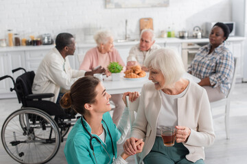 Positive nurse looking at patient with tea near blurred elderly people in nursing home
