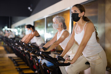 Fototapeta na wymiar Young girl in face mask warming up on training using exercise bike in gym