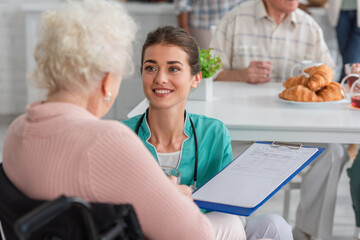 Smiling nurse holding clipboard near blurred senior woman in wheelchair in nursing home