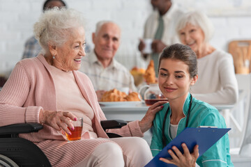 Smiling senior woman holding tea in wheelchair near nurse with clipboard in nursing home