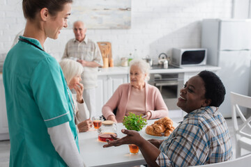 Smiling african american woman talking to nurse near tea and croissants in nursing home