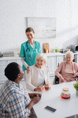 Smiling nurse standing near multiethnic patients with smartphones and tea in kitchen of nursing home