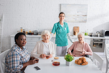 Young nurse standing near interracial elderly women with devices and tea in nursing home