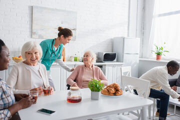 Nurse talking to elderly woman in wheelchair near interracial people in nursing home