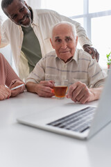 Smiling african american man hugging senior friends with tea near laptop in nursing home