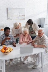 Positive multiethnic senior people using laptop near tea and fruits in kitchen