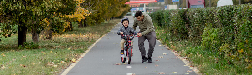 Father teaches his son to ride a bicycle on the bike path in the park. The father is holding a...