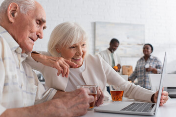 Obraz premium Positive elderly woman pointing at laptop near friend with cup of tea in nursing home