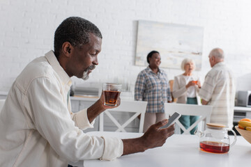 Smiling senior african american man with tea using smartphone in nursing home