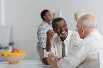 Smiling african american man showing yes gesture near senior friend with smartphone and blurred women in nursing home