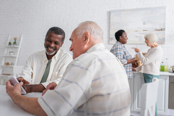 Obraz premium Cheerful senior men using smartphone near blurred multiethnic friends in nursing home