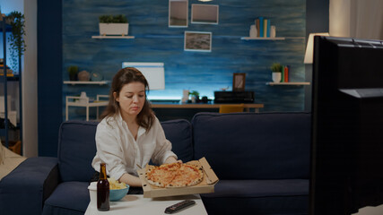 Person bringing box of pizza from delivery man on table in living room. Young woman with fast food and alcoholic beverage sitting on sofa watching television. Adult ordering takeaway meal