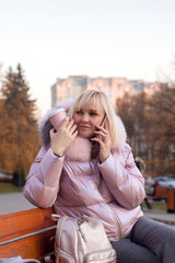 A young woman in stylish warm clothes and a cap sits on a bench against the background of the street and park, drinks coffee, tea and talks on the phone