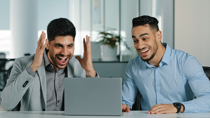Two Arab young male businessmen sitting in modern office using laptop employees satisfied with result of negotiations, having concluded contract, Indian colleagues receiving good news feeling thankful