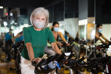 Obraz na płótnie Canvas Portrait of active mature woman wearing face mask for disease protection training on stationary bike workout in gym