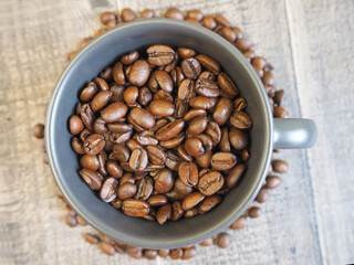 a dark gray mug filled with roasted whole coffee beans stands on a wooden background with coffee beans at the bottom of the mug