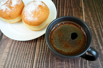 two doughnuts, sprinkled with powdered sugar, lay on a white plate next to a dark coffee mug on a wooden table. side view