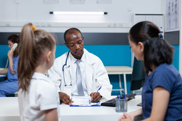 Fototapeta premium African american pediatrician doctor writing sickness symptoms on clipboard discussing medical treatment with family during clinical examination in hospital office. Health care treatment