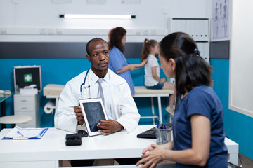 Fototapeta premium African american pediatrician doctor holding tablet computer with kid radiography on screen discussing healthcare treatment with patient mother during consultation in hospital office. Medicine service