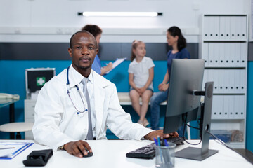 Fototapeta premium Portrait of african american practitioner doctor sitting at desk in hospital office analyzing sickness expertise on computer during clinical appointment. Pediatric man typing illness symptoms