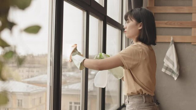 Medium slowmo shot of young Asian woman in casualwear and rubber gloves cleaning windows at home during housework, using cleaner spray and cloth