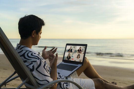 Asian Business Man Having Remote Video Conference Call With His Business Team At The Beach During Vacation In Holiday..