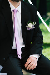 Groom sit wearing black suit outfit with white rose boutonnieres and light purple tie on white shirt outdoors on wedding - 471453773