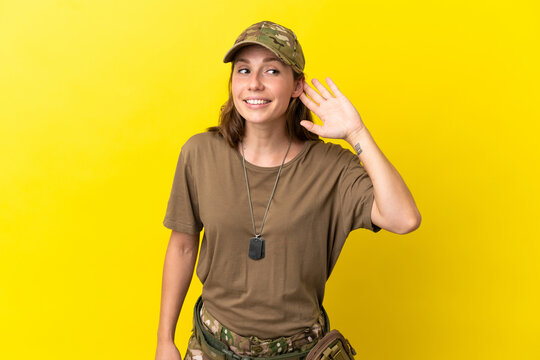Military caucasian woman with dog tag isolated on yellow background listening to something by putting hand on the ear