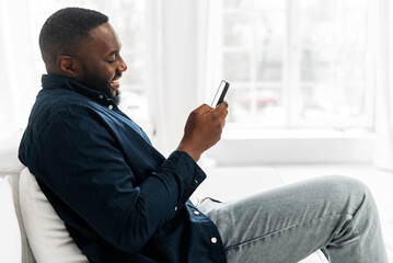 Smiling young African-American holding smartphone sitting on sofa at modern apartment and chatting online, enjoying texting in dating app, answering messages, writes a post for social media, side view