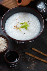 Congee or a type of asian rice porridge served in a black bowl, middle closeup, vertical shot on a brown stone surface