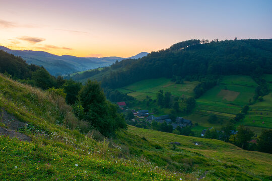rural landscape in mountains at dawn. beautiful carpathian nature scenery with grassy fields and meadows between forested hills. village in the distant valley. idyllic alpine countryside at twilight