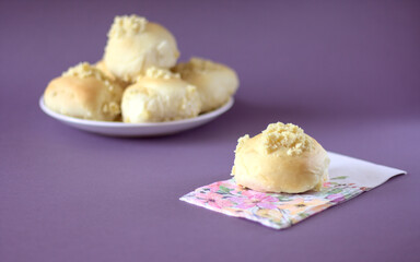 Homemade yeast roll (bun) with crumble on a flowery napkin. Pleasant gray - violet background, in the distance a plate with buns. Copy space.