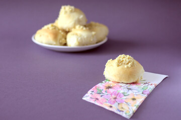 Homemade yeast roll (bun) with crumble on a flowery napkin. Pleasant gray - violet background, in the distance a plate with buns. Copy space.