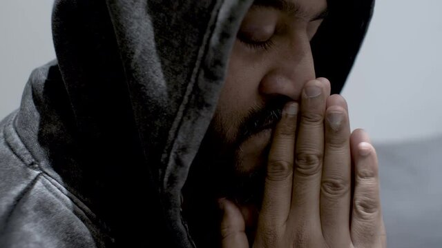 A close up shot of an Indian man holding his hands in front of his mouth, eyes closed as he rocks back and forth wrestling with his deep dark anxious thoughts