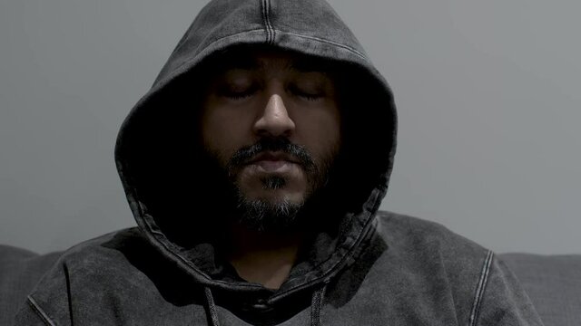 A close up shot of the face of an Indian man wearing a hoodie, dollying out as he remains motionless with his eyes closed as he calmly meditates in complete silence