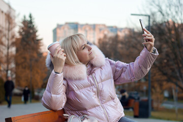 Attractive young woman blonde girl makes a selfie on a bench on the background of the street, the blogger communicates in video chat, the tourist is resting and drinking coffee in the park