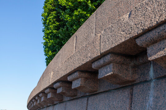Close-up of a granite architectural detail: a curved cornice with stepped modillions, set against a blue sky and lush greenery. Highlights the texture and craftsmanship of historic stonework.