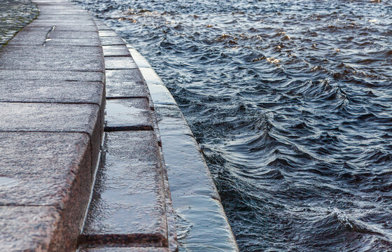 Close-up of a stone embankment meeting turbulent water: rectangular granite slabs form a sturdy walkway, while the churning waves reveal the raw force of the river. The contrast between the rigid stru