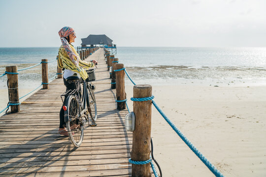Portrait of a smiling woman dressed in light summer clothes, sunglasses with bicycle on the wooden sea pier on the sandy Zanzibar beach.Careless vacation in the tropical countries concept image