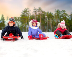 childhood, leisure and season concept - group of happy little kids sliding down snow hill on sleds in winter over forest or park background