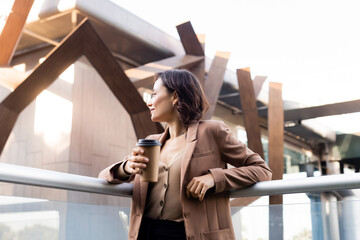 Fototapeta premium brunette woman in beige blazer looking away on balcony of building outdoors