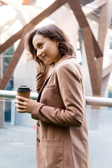 smiling woman in beige blazer holding coffee to go outdoors