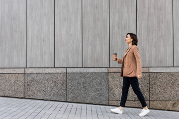 side view of stylish woman walking with coffee to go along grey wall