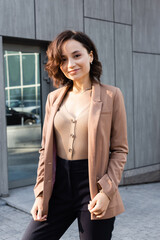 brunette woman in beige blazer smiling at camera while standing outdoors