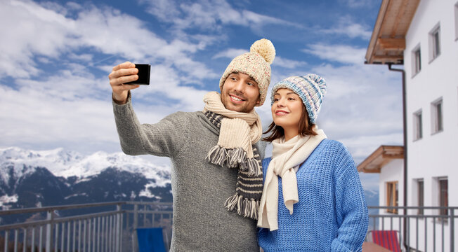 travel, tourism and winter holidays concept - happy couple in knitted hats and scarves taking selfie by smartphone over mountains and ski resort background
