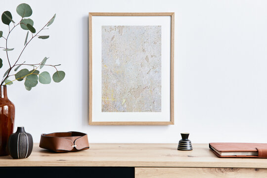 Stylish interior of living room with mock up poster frame, wooden commode, book, eucalyptus leaf in ceramic vase and elegant personal accessories. Minimalist concept of home decor. Template.