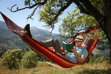 Obraz na płótnie Canvas Handsome man reading book in hammock outdoors on sunny day