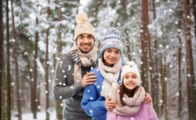 family, christmas and holidays concept - happy mother, father and little daughter in knitted hats and scarves over winter forest background