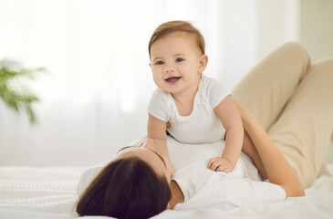 Obraz na płótnie Canvas Little baby girl and her mom in morning having fun together playing on white bed. Smiling happy baby lying on recognizable mom in bright bedroom or nursery. Concept of motherhood and childhood.