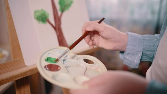 Teenager girl paints tree on paper at home. Close-up of palette, selective focus.